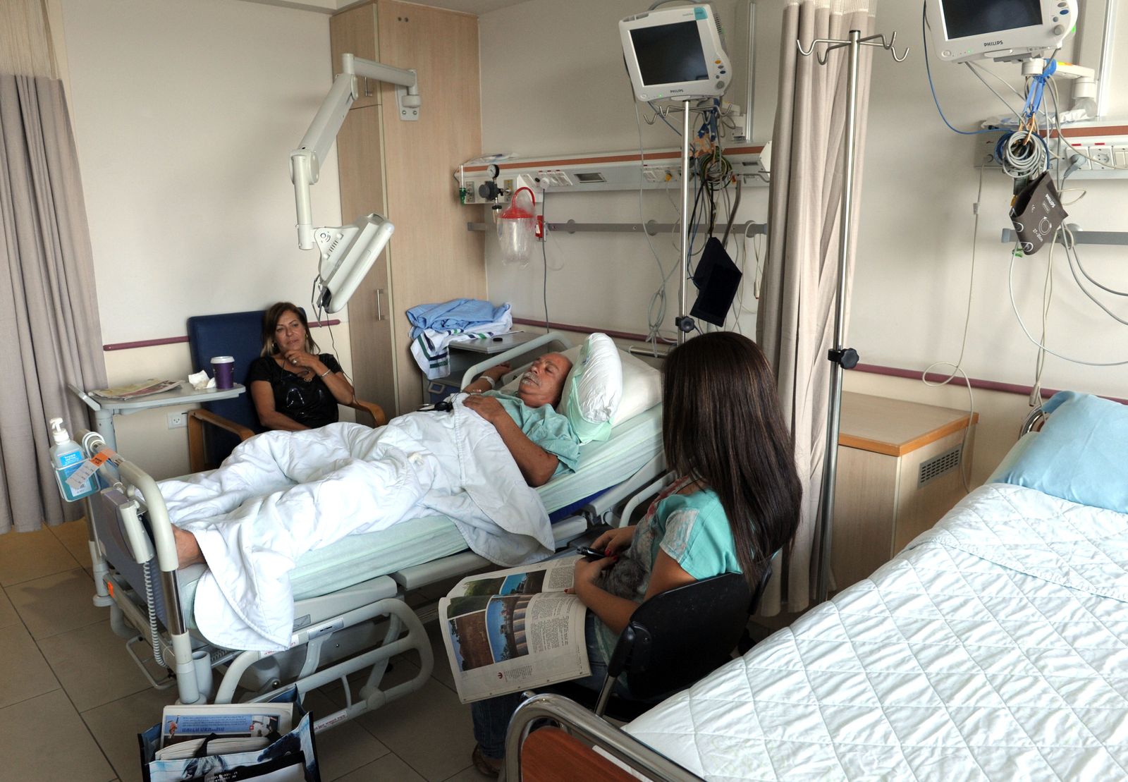 A patient resting in a hospital bed while two family members sit beside him.