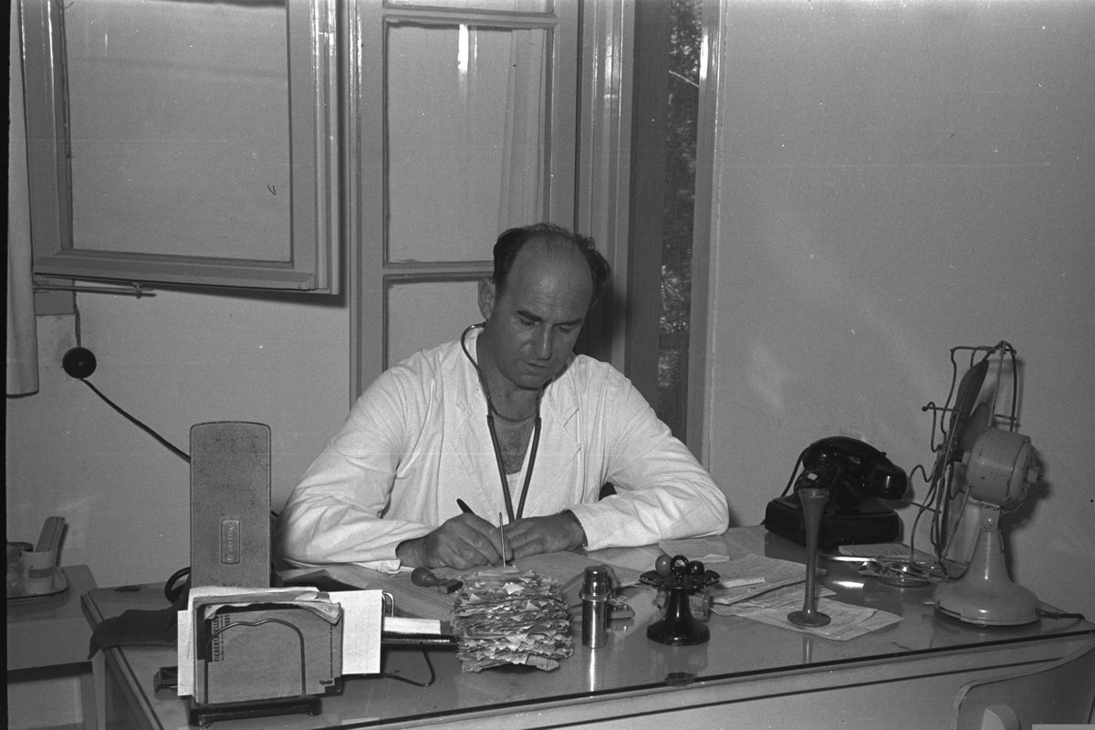 A doctor in a white coat writing at his desk in a clinic.