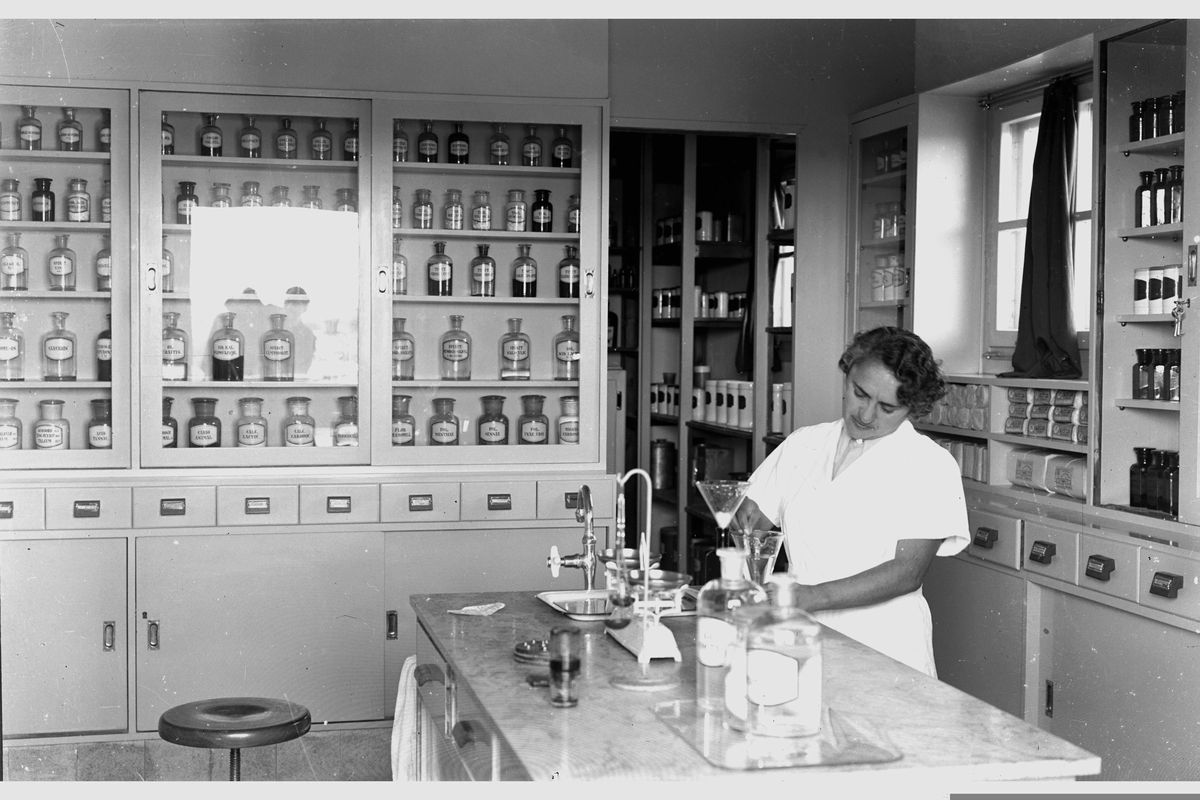 A pharmacist at a counter in front of shelves of medicine jars.