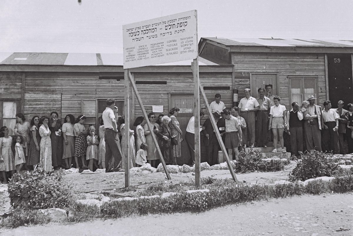 A long line of people outside wooden huts, under a sign for the health fund.
